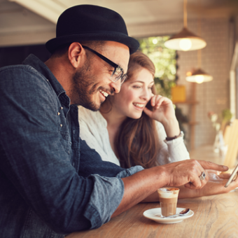 Ein junger Mann und eine junge Frau sitzen in einem Kaffeehaus und schauen sich etwas auf einem Smartphone an.