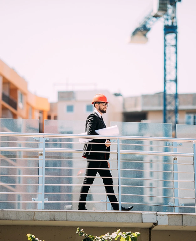 Ein Mann im Anzug und mit Bauhelm geht mit einem Plan unter dem Arm auf einer Großbaustelle