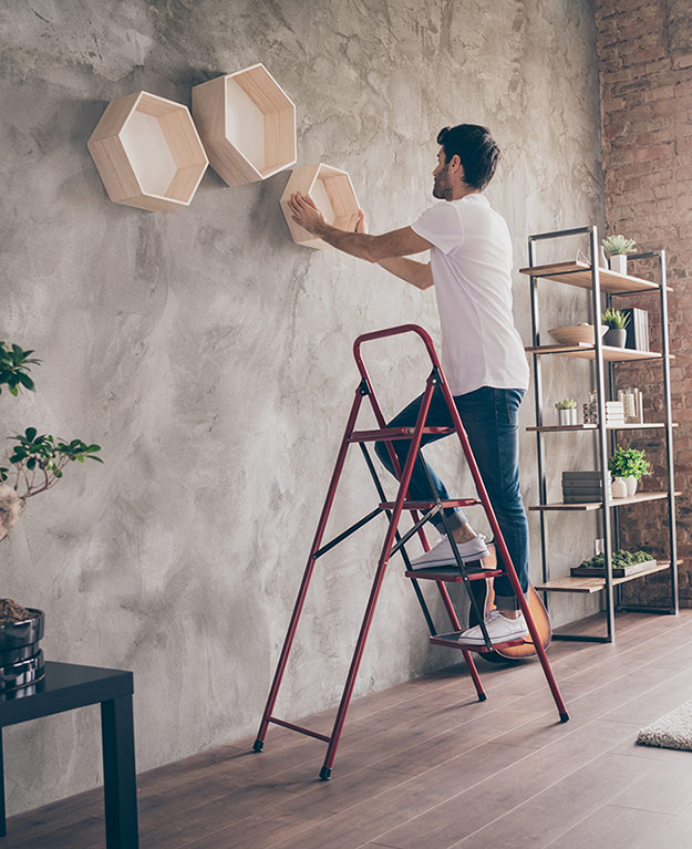 Mann steht auf einer Leiter im Wohnzimmer und montiert Holzboxen an der Wand