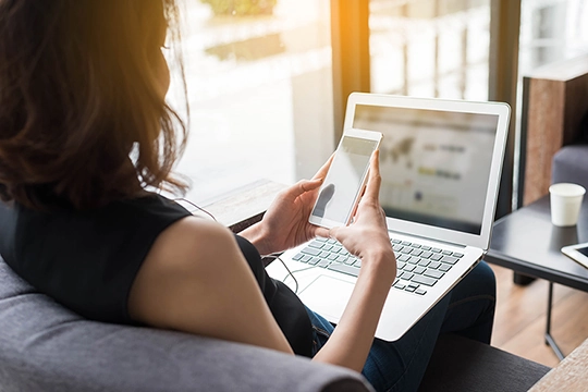 Frau sitzt auf einem Sofa und schaut auf ihr Handy und ihren Computer
